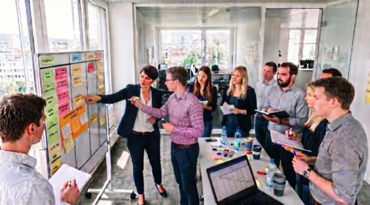 Ein Projektteam versammelt sich in einem Schweizer Büro um eine große Aufgabenwand, während eine Person eine Aufgabe einem Teammitglied zuweist.